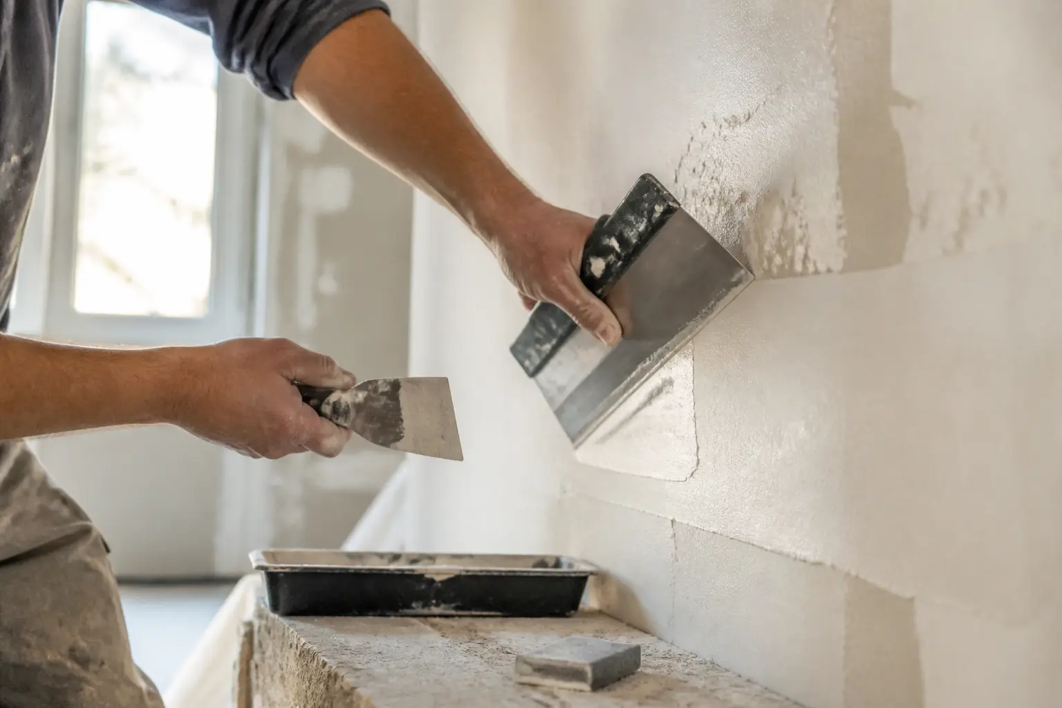 Lisser un enduit sur un mur intérieur avec couteau à enduire, lame large, zones réparées et lumière rasante.