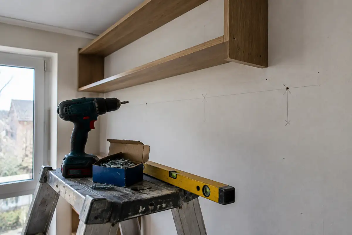 Perceuse, chevilles métalliques et niveau posés sur un escabeau sous une étagère haute en cours de pose sur une cloison