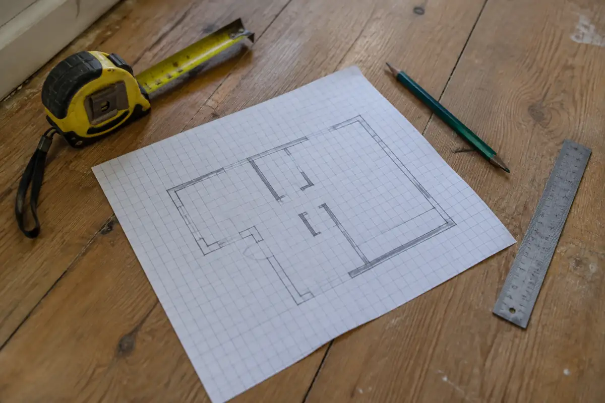 Plan de pièce dessiné sur papier quadrillé, avec un mètre ruban, un crayon et une règle posés sur un parquet